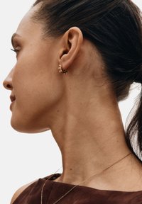 Kuldsetest rõngaskõrvarõngastest koosnevad kõrvarõngad, millel on naelu meenutavad aksendid ja tekstuurne viimistlus, kantud sirge juukseprofiiliga inimese kõrvas, näidates külgvaadet.