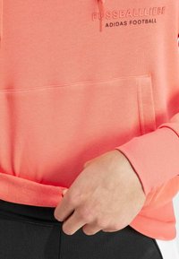 Koraal hoodie gemaakt van zachte stof, met een voorzak in kangoeroestijl, ribgebreide manchetten en geborduurde tekst "FUSSBALLLIEBE" en "ADIDAS FOOTBALL."