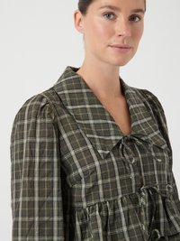 Žena u zelenoj kariranoj haljini s velikim ovratnikom, napučenim rukavima i vezanim mašnama na vratu i struku, na jednobojnoj bijeloj pozadini.