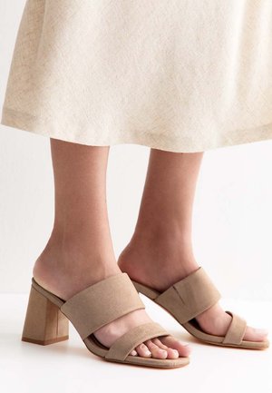 Piedi che indossano sandali beige con tacco a blocco e due ampie fasce, abbinati a una gonna chiara di lunghezza media.