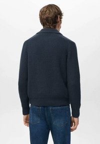 Granatowy sweter dziany o prążkowanej fakturze, z kołnierzem szalowym i dopasowanym dołem. Noszony z niebieskimi dżinsami, widok od tyłu.