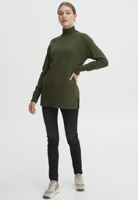 Olivgrön polotröja i mjukt stickat material med långa ärmar, sidofickor och en liten logodetalj på fållen. Matchad med svarta jeans och sneakers.