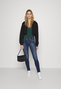 Hoodie cropped preto, blusa verde, jeans skinny azuis, cinto preto com acabamentos em metal, segurando uma bolsa preta, ténis brancos. Fundo simples.