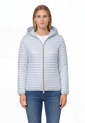 Donna che indossa una giacca trapuntata grigio chiaro con cappuccio e cerniera frontale, e jeans blu, in piedi davanti a uno sfondo bianco.