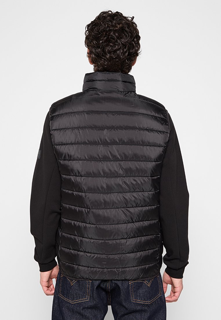 Gilet imbottito nero con design a coste orizzontali, colletto alto e chiusura con zip. Abbinato a una maglietta nera a maniche lunghe e pantaloni di jeans.