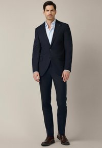 Granatowy garnitur z dopasowaną marynarką, pasującymi spodniami i jasnoniebieską koszulą. Całość dopełniają brązowe skórzane buty eleganckie.