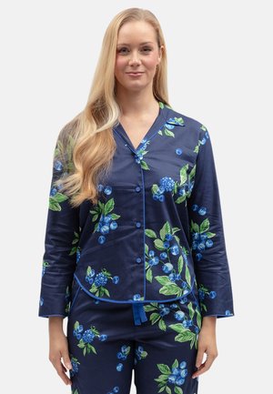 Frau mit langen blonden Haaren, die marineblaue Pyjamas mit blau-grünem Blumenmuster trägt, steht vor einem einfarbigen hellen Hintergrund.