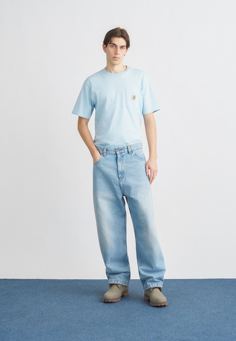 Hellblaues Baumwoll-T-Shirt mit Tasche, kombiniert mit locker sitzenden hellen Jeans und grauen Stiefeletten, stehend auf einer blau Teppichoberfläche.