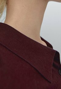 Bordeauxkleurige trui met een gebreide textuur, voorzien van een kraag en een knoopdetail bij de hals, met een close-up van de stof en huid.
