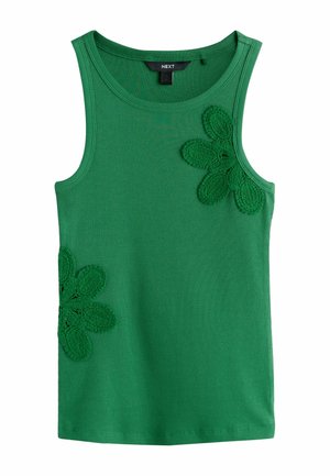 Žena stojí a má na sebe zelený tielko bez rukávov s kvetinovými detailmi, voľné čierne nohavice s opaskom a čierne žabky.