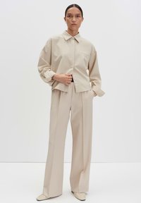 Donna con capelli scuri, indossa una camicia beige oversize con bottoni, pantaloni a gamba larga e scarpe a punta coordinate, in piedi su uno sfondo bianco.