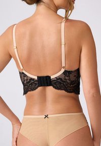 Kobieta w beżowym i czarnym koronkowym biustonoszu z regulowanymi ramiączkami oraz beżowych majtkach z małą czarną kokardką na tylnej górze.