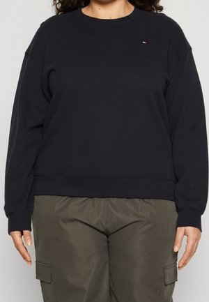Person iført en sort langærmet sweatshirt med et lille logo på brystet og olivengrønne cargobukser med lommer.