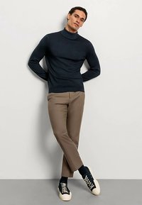 Marineblauer Rollkragenpullover kombiniert mit hellbrauner Hose und marineblauen Sneakers. Das Material wirkt weich; klare Linien und minimalistisches Design.