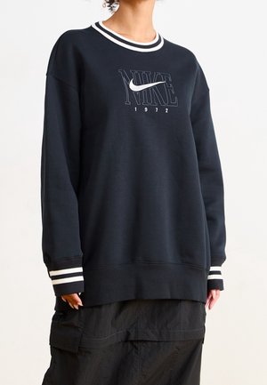 Person iført en sort Nike-sweatshirt med hvide stribede ærmekanter og krave, der viser "NIKE"-logoet og "1972" på brystet.
