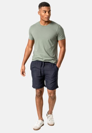 Mann geht, trägt ein hellgrünes kurzärmeliges T-Shirt, marineblaue Shorts, weiße Turnschuhe, eine Hand in der Tasche, schaut zur Seite.