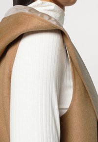 Couche extérieure beige en tissu doux avec une capuche, portée sur un haut blanc côtelé à col haut. Texture lisse et silhouette épurée.