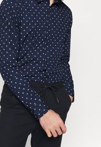 Camisa de botonadura azul marino con un pequeño patrón geométrico blanco; combinada con pantalones negros con cordón. Tela suave y diseño ajustado.