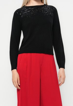 Femme portant un pull noir à manches longues avec des sequins noirs au niveau du col et un pantalon large rouge vif.