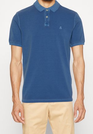 Mężczyzna w niebieskiej koszulce polo z krótkim rękawem i dwoma guzikami oraz beżowych spodniach, stojący na jednolitym tle.