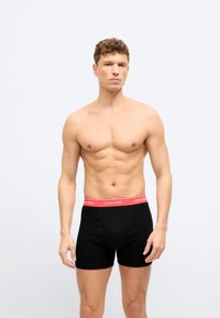 Άνδρας χωρίς πουκάμισο με σγουρά μαλλιά, φορώντας μαύρα ανδρικά εσώρουχα με κόκκινη μέση Calvin Klein, στέκεται μπροστά σε ένα απλό φωτεινό φόντο.