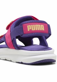 PUMA-sandaalit ovat violetin ja vaaleanpunaisen väriset, joissa on teksturoitu hihna, säädettävä suljenta ja pehmustettu valkoinen pohja. Huomaa merkkilogolla varustettu takapuoli.