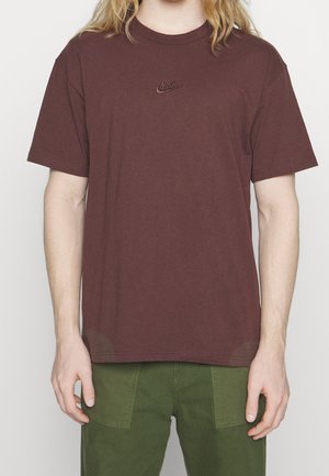 Osoba nosząca gładki brązowy t-shirt Nike z krótkimi rękawami i oliwkowe spodnie, pokazana od ramion do talii.