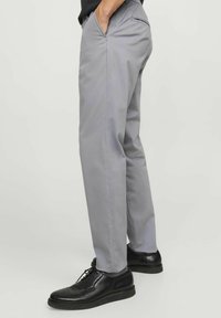 Uomo che indossa pantaloni dritti grigio chiaro e scarpe eleganti in pelle nera, in piedi con una mano in tasca contro uno sfondo semplice.