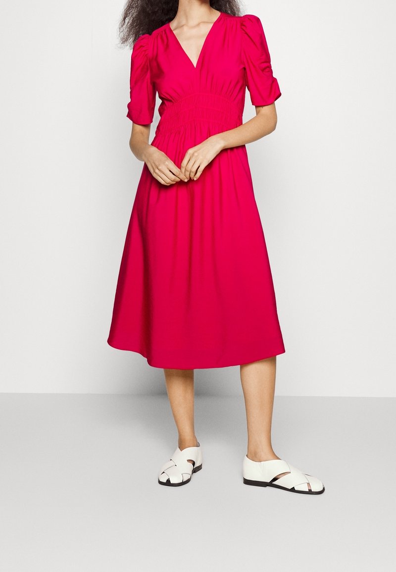 Femme portant une robe midi rose vif à manches bouffantes et des sandales blanches plates à bouts fermés, debout devant un fond clair uni.