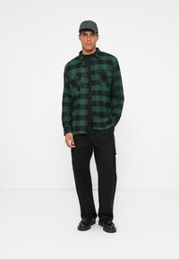 Camicia di flanella a quadri verdi e neri con due tasche frontali, abbinata a pantaloni cargo neri e scarpe nere, completata da un cappellino nero.