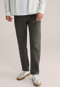 Man in een donkere, grijze recht-toe-recht-aan broek, met witte en beige sneakers, en een wit shirt met daaroverheen een gestreepte jas.