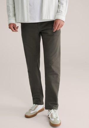 Man in een donkere, grijze recht-toe-recht-aan broek, met witte en beige sneakers, en een wit shirt met daaroverheen een gestreepte jas.
