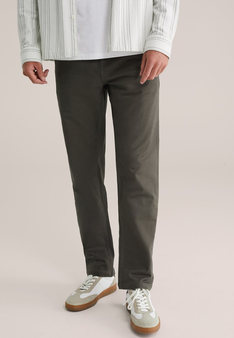 Man in een donkere, grijze recht-toe-recht-aan broek, met witte en beige sneakers, en een wit shirt met daaroverheen een gestreepte jas.