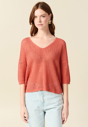 Giovane donna che indossa un maglione corallo a maglia aperta con scollo a V, maniche fino al gomito e jeans a vita alta azzurri chiaro, su sfondo semplice.