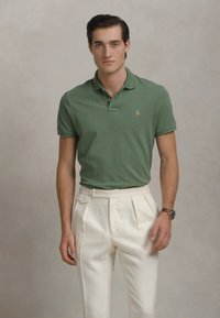 Mlad moški v zeleni polo majici in belih hlačah s visokim pasom in gubami stoji pred nevtralno ozadjem.