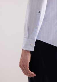 Camicia a righe blu chiaro con polsini abbottonati a due bottoni e un piccolo logo ricamato a forma di rosa sulla manica. Texture liscia.