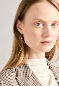 Silberne Creole Ohrringe mit klaren Edelsteinen, die eine glatte, glänzende Oberfläche aufweisen, getragen von einer Person mit blonden Haaren und einem gemusterten Blazer.