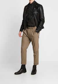 Giacca di pelle nera, camicia nera con pois crema, pantaloni khaki a pieghe e stivaletti neri con una finitura liscia, su uno sfondo semplice.