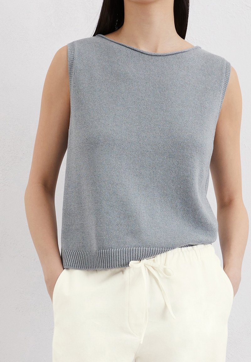 Haut en maille gris sans manches porté avec un pantalon blanc taille haute à cordon, mains dans les poches, sur un fond clair uni.