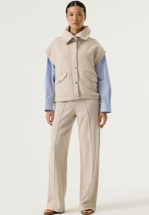 Chaqueta corta beige con gran cuello y bolsillos delanteros, combinada con pantalones beige de corte holgado con costuras laterales. Textura de tela suave.