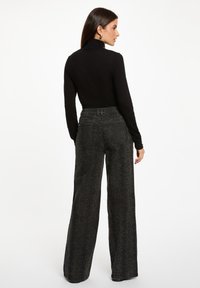 Schwarzer Rollkragenpullover kombiniert mit hochgeschnittenen, ausgestellten Hosen aus einem strukturierten, dunkel gemusterten Stoff. Die Rückansicht zeigt die taillierten Ärmel und den Bund.