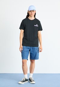 Tricou din bumbac negru cu grafică roz și alb, pantaloni scurți din denim albastru, șosete albe, adidași negri și albi, și o șapcă albastră deschisă.