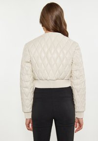 Giacca bomber trapuntata beige con polsini e orlo a coste. Tessuto liscio con motivo a diamante. Indossata sopra pantaloni neri aderenti.
