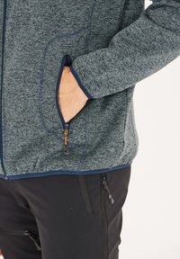 Grå fleecetröja med marinblå detaljer, med en dragkedjefickor och hög krage. Bärs över svarta byxor med flera dragkedjefickor.