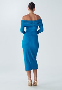 Robe bleue à épaules dénudées, manches longues, tissu texturé, silhouette ajustée, longueur mi-mollet, fermeture éclair au dos, portée avec des talons à brides.