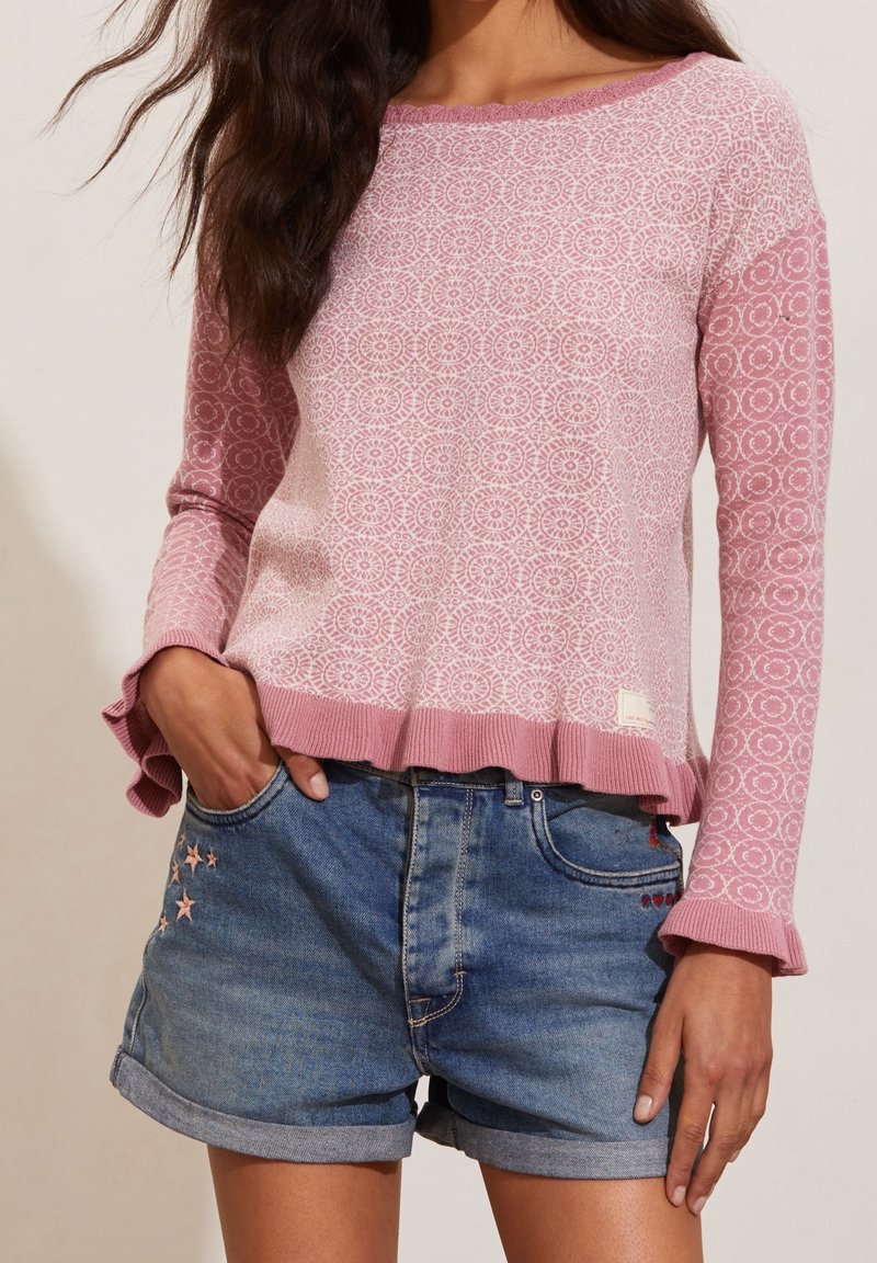 Femme portant un pull à manches longues rose à motifs avec des poignets et un ourlet volantés, associé à un short en denim bleu avec des broderies d'étoiles.
