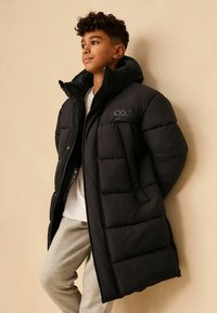 Čierny páperový kabát s vysokým golierom, s prešívaným dizajnom a predným zipsom. Sú vidno aj svetlošedé nohavice a biele tričko.