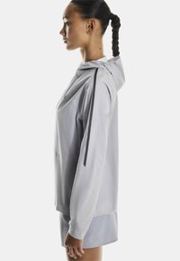 Svetlo siva športna jakna iz gladkega materiala, ki ima kapuco, črne črte na rokavih in sproščen kroj.