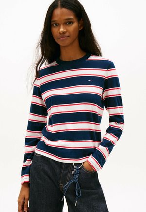 Kvinde iført en navy- og hvide stribet langærmet skjorte med røde detaljer og mørke jeans, stående med den ene hånd i lommen og en flettet nøglering hængende fast.