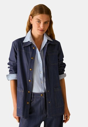 Donna che indossa una giacca e pantaloni di jeans blu scuro con le maniche arrotolate sopra una camicia azzurra con bottoni, in piedi davanti a uno sfondo neutro.